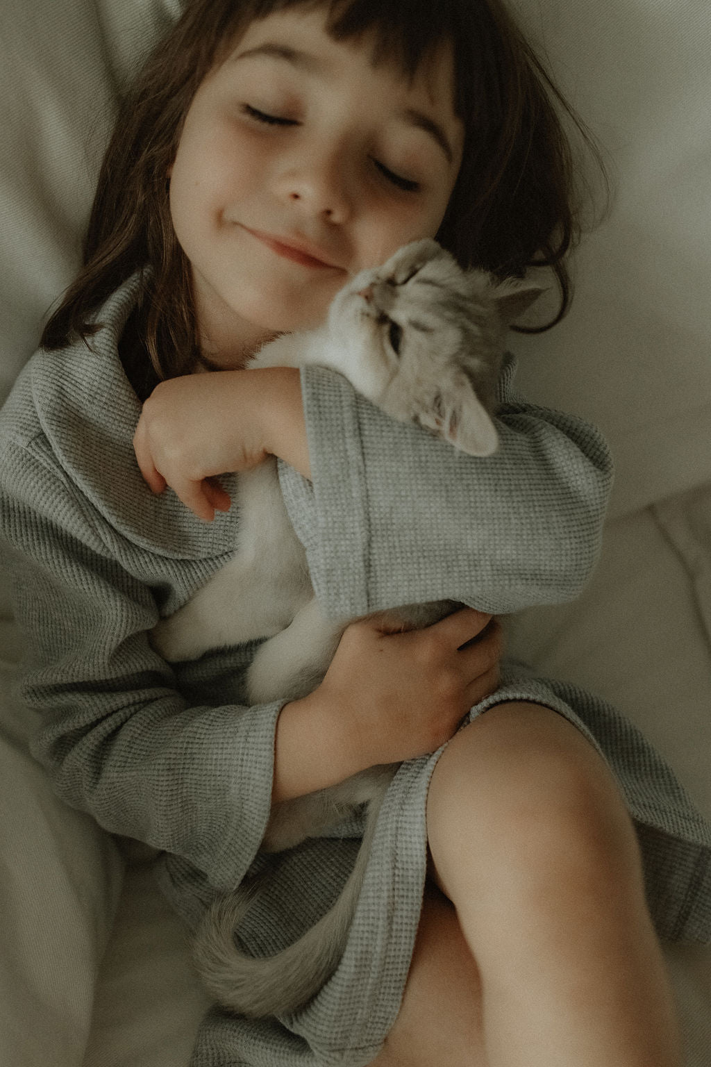 Child in a gray robe holding a cat, lying on a soft surface.