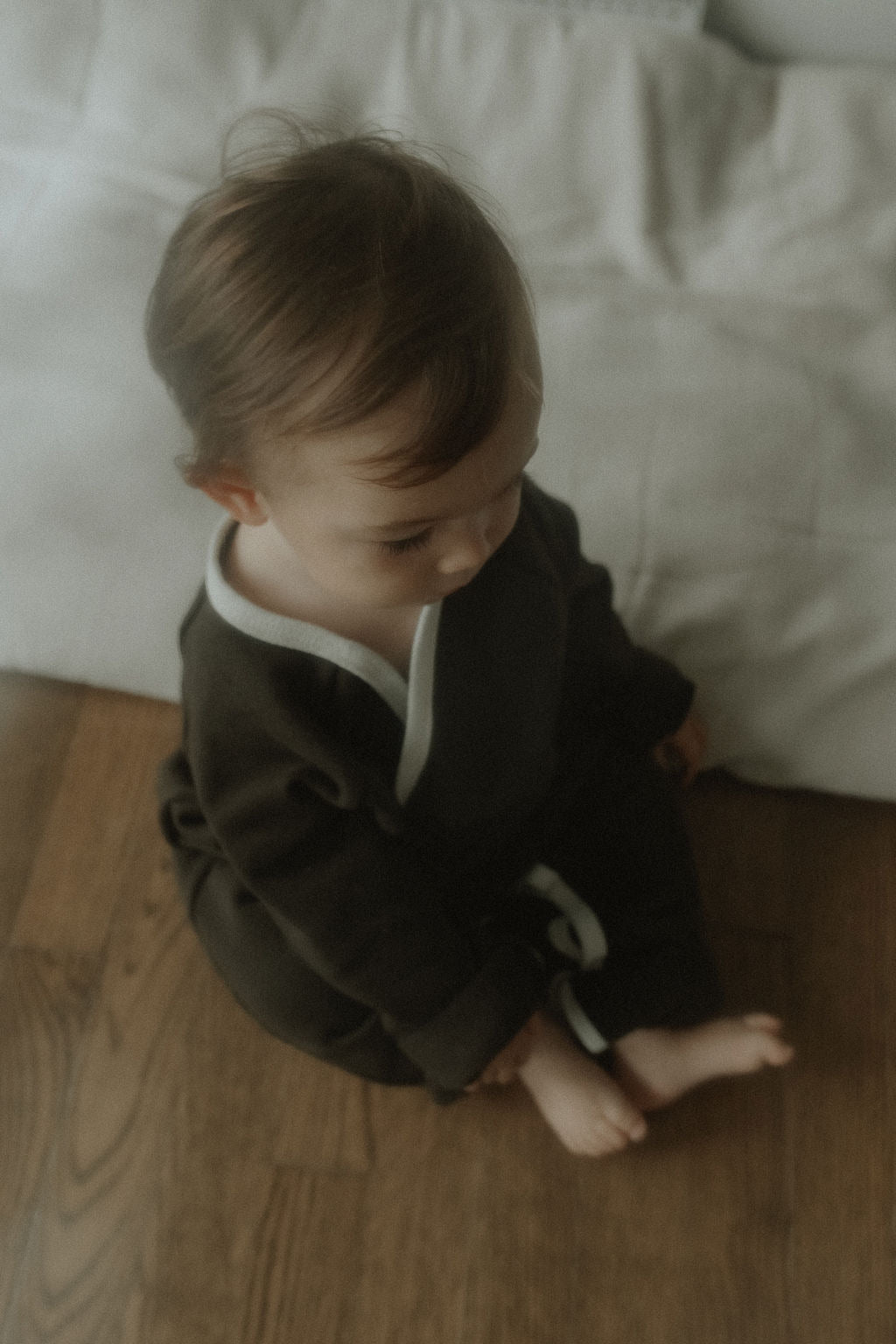 Child in a dark outfit sitting on a wooden floor with light-colored cushions in the background