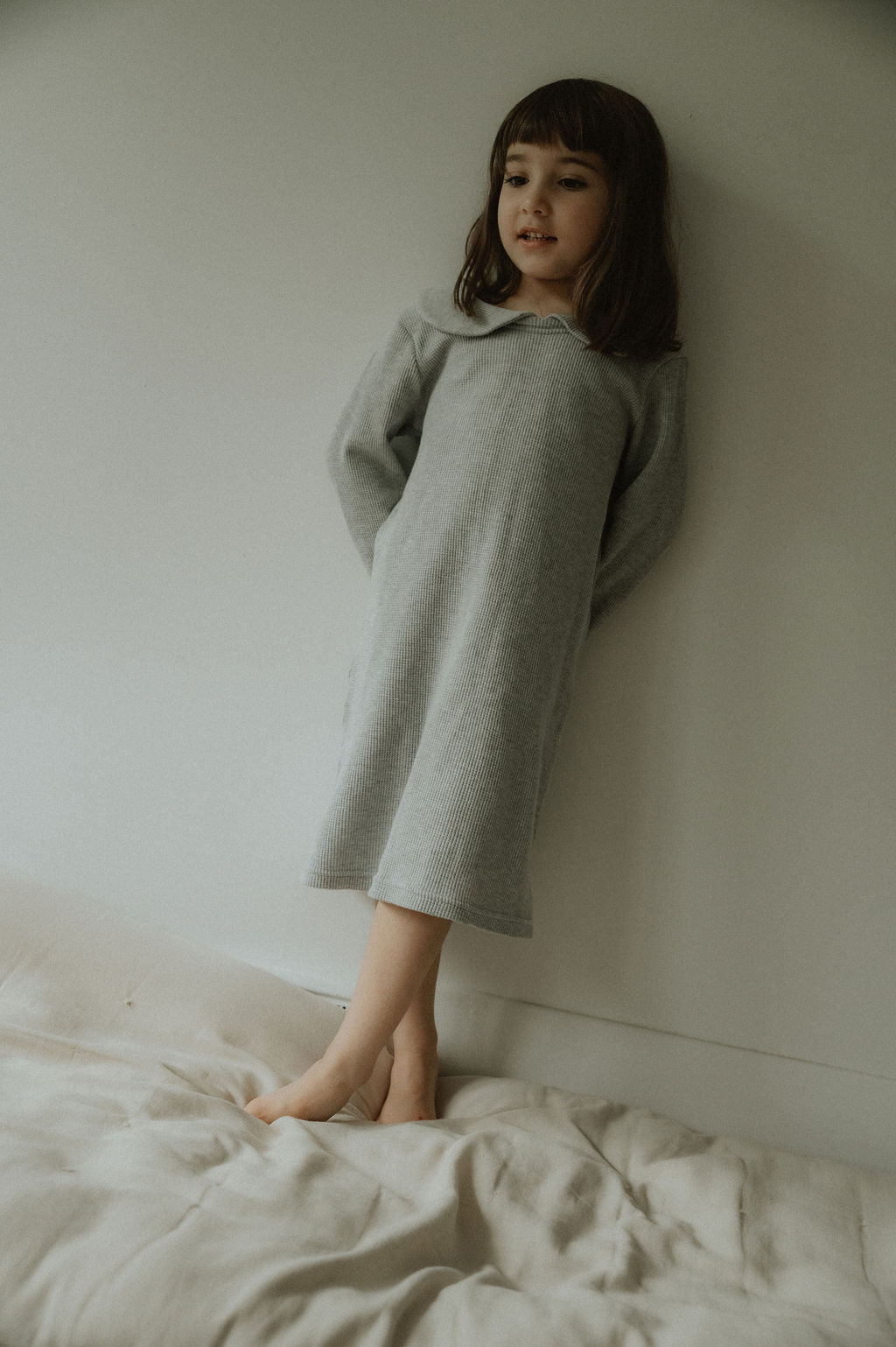 Young girl in a gray dress standing on a bed against a plain wall