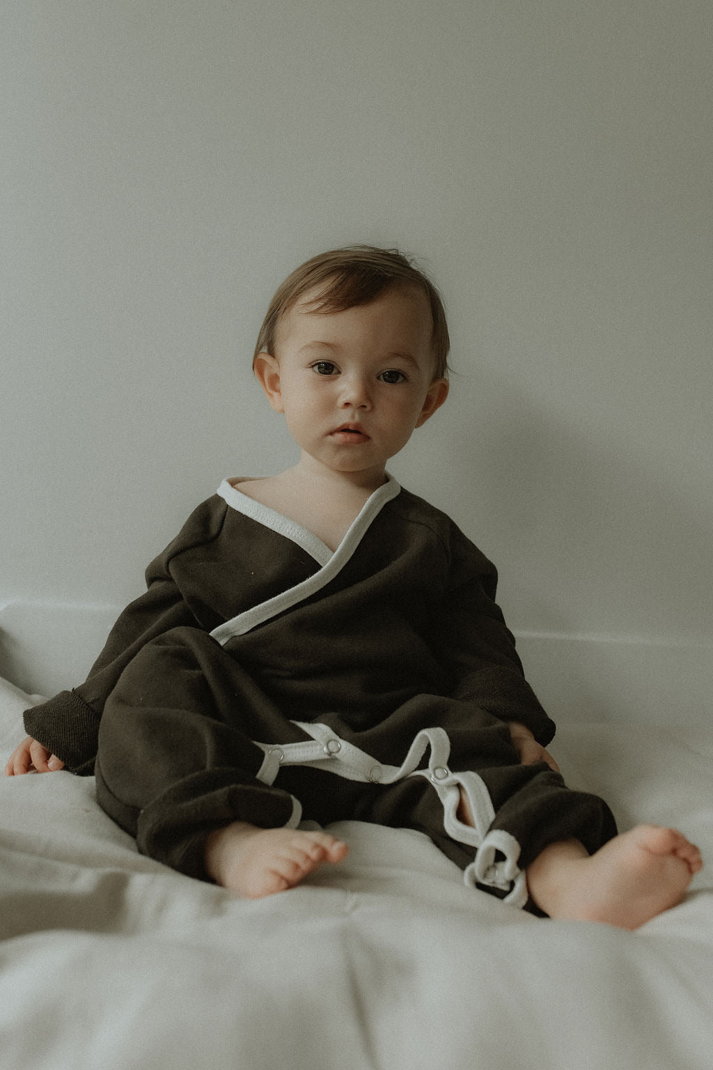 Baby wearing a dark brown outfit with white trim sitting on a light-colored surface.