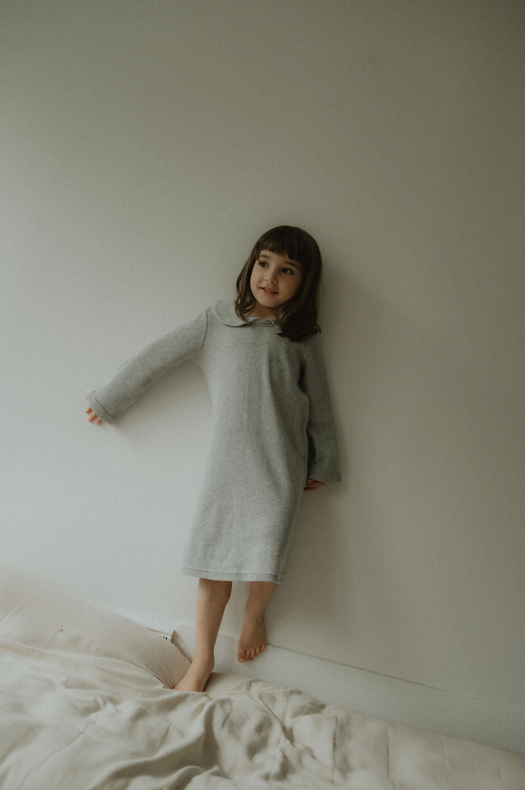 Child in a gray dress standing against a plain wall with a bed in the foreground.