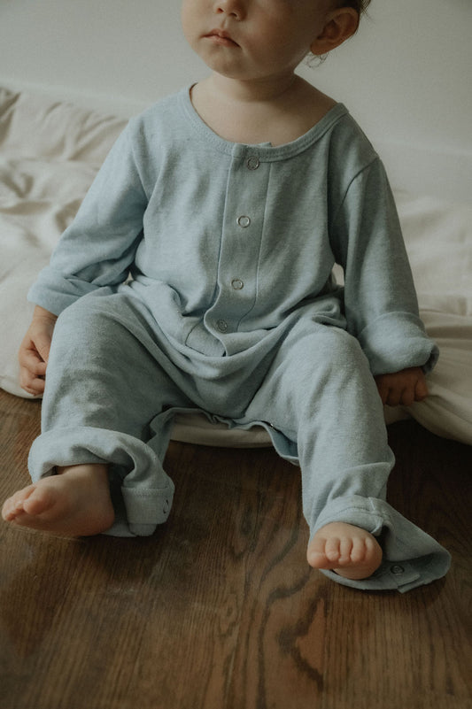 Child wearing a light blue onesie sitting on a wooden floor.